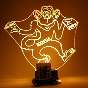 Może przedstawiać: Żółta, podświetlana nocna lampka akrylowa w kształcie małpy z napisem "Rochelle". Lampka nocna ma wielokolorową podstawę LED.