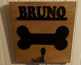 Op de afbeelding: Een houten hondenriemhouder met een zwart botontwerp en de naam "BRUNO" in zwarte letters. De houder heeft een metalen haak om een riem aan te hangen.