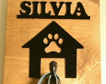 May include: A wooden sign with a black metal dog house cutout featuring a paw print and a hook for a leash. The sign reads "SILVIA" at the top.