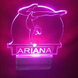 May include: A pink LED night light featuring a gymnast in a handstand pose. The name "ARIANA" is displayed in white block letters on the base of the light. The light emits a soft, ambient glow, perfect for a child's room.