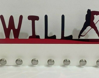 Può includere: Un portachiavi bianco con il nome "WILL" in lettere rosse e nere e una silhouette di un giocatore di hockey. Il portachiavi ha otto ganci argentati.