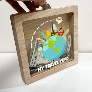 May include: Wooden piggy bank with a clear front panel. The panel shows a globe with a blue and green design, surrounded by images of famous landmarks. The text "MY TRAVEL FUND" is printed below the globe.