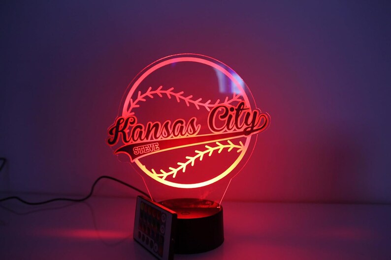 May include: A baseball-themed LED lamp with the words "Kansas City" in red script, and "STEVE" below. The lamp is illuminated with a red glow, and has a remote control.