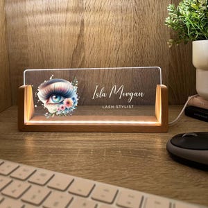 May include: A lighted wooden desk sign with the name "Isla Morgan" and the title "Lash Stylist" printed on a clear acrylic panel. The panel features a watercolor eye design with floral accents. The sign is illuminated by a warm light.