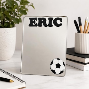 以下が含まれることがあります： 上部に太い黒文字でERICという名前が入り、右下に白黒のサッカーボールのグラフィックが描かれた長方形の鏡。鏡にはシンプルなフレームが付いており、表面は反射するようになっています。