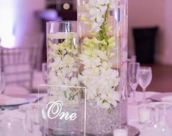 May include: A clear glass vase filled with white orchids and water, with a clear acrylic table number sign that says "One" on a black base. The vase is sitting on a white tablecloth with other clear glass vases and candles.