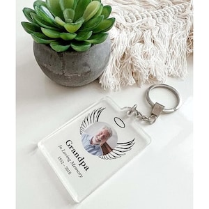 May include: Clear acrylic keychain with a photo of a smiling man, wings, and a halo. Text reads "Grandpa In Loving Memory 1952-2018." The keychain has a silver metal keyring attached. A small succulent in a gray pot is in the background.