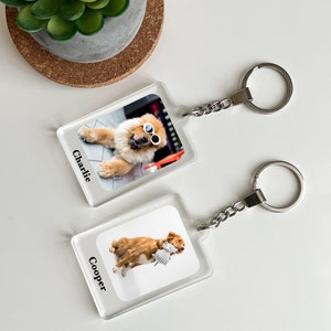 May include: Two clear acrylic keychains with dog photos. The top keychain features a Chow Chow wearing sunglasses with the name "Charlie" printed below. The bottom keychain features a golden retriever holding a stuffed toy with the name "Cooper" printed below.