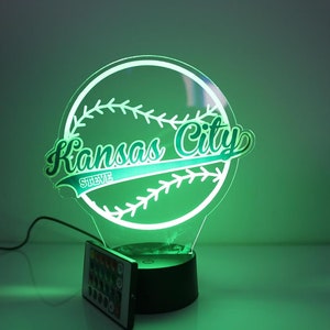 May include: A glowing baseball-themed night light with the words "Kansas City" and the name "Steve" inscribed. The light is green and has a remote control. The design is a baseball with stitching.