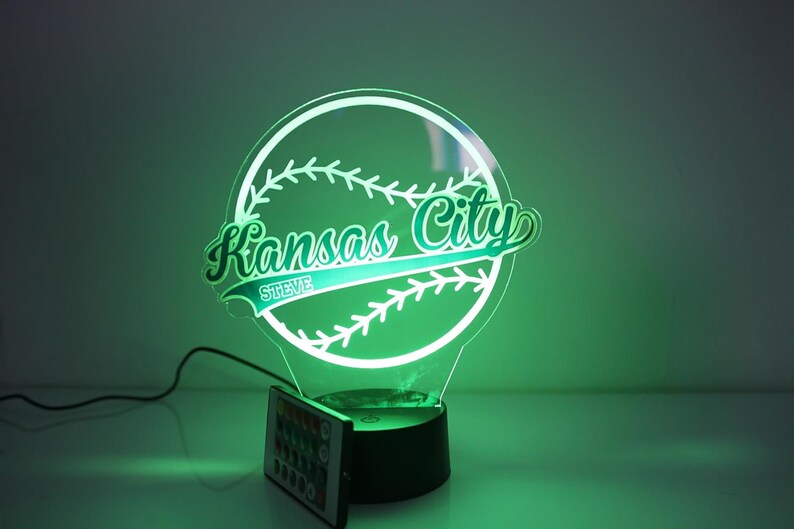 May include: A glowing baseball-themed night light with the words "Kansas City" and the name "Steve" inscribed. The light is green and has a remote control. The design is a baseball with stitching.
