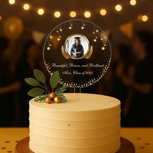 Könnte beinhalten: Ein Tortenaufsatz zur Graduierung mit einem Foto einer Absolventin in Robe und Hut. Der klare Acryl-Topper hat goldene Akzente und die Aufschrift "Beautiful, Brave, and Brilliant, Ava, Class of 2025." Der Kuchen ist hellgelb mit einem dekorativen Blatt- und Beerenarrangement.