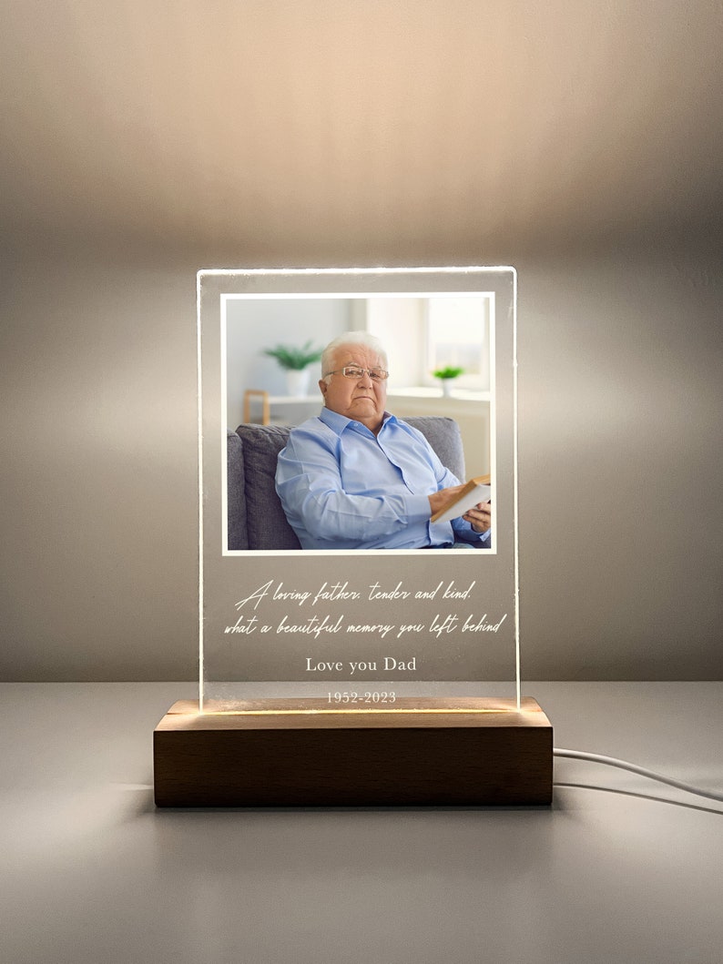 May include: A clear acrylic photo frame with a wooden base, displaying a black and white photo of a man. The frame has a white inscription that reads "A loving father, tender and kind. What a beautiful memory you left behind. Love you Dad 1952-2023."
