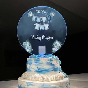 May include: A round, illuminated acrylic cake topper featuring the text Oh Boy and Baby Mason, decorated with blue floral accents and a graphic of baby clothes hanging on a line. The topper is placed on a two-tiered cake with blue and white frosting.