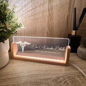 Può includere: Un'insegna da scrivania illuminata con base in legno. Il pannello in acrilico trasparente mostra il simbolo del Caduceo, il nome "Dr. Amanda Blake" e "DOCTOR OF PHARMACY" in caratteri bianchi. L'insegna è illuminata da una luce calda.