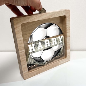 May include: Wooden piggy bank with a clear front panel. The front panel features a black and white soccer ball with the name "HARRY" printed across the ball. The piggy bank has a slot on the top for coins.