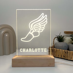 Puede incluir: Una luz nocturna de acrílico blanco con un diseño de zapatilla de correr y ala. El nombre "CHARLOTTE" está impreso debajo del diseño. La luz está sobre una base de madera.