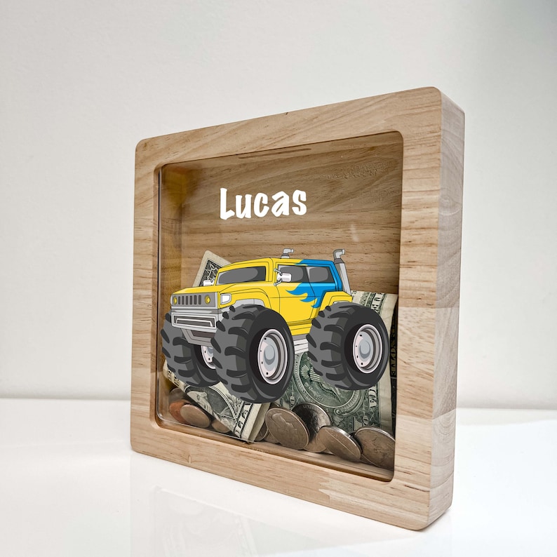 May include: A wooden money box with a clear front panel. The box has a yellow monster truck graphic and the name "Lucas" printed on the front. The box is filled with coins and bills.