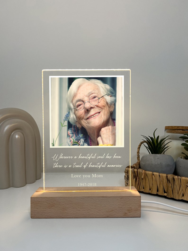 May include: A white acrylic light-up photo frame with a picture of an older woman smiling. The frame has the text "Wherever a beautiful soul has been there is a trail of beautiful memories" and "Love you Mom 1947-2018".