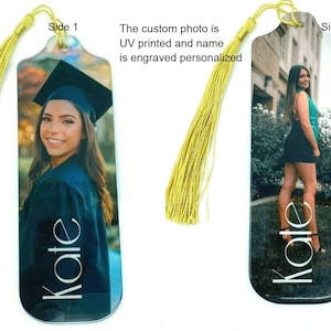 May include: A personalized glass bookmark with a tassel. The bookmark features a photo of a person in a graduation cap and gown on one side and a photo of the same person in a black dress on the other side. The name "Kate" is engraved on both sides of the bookmark.