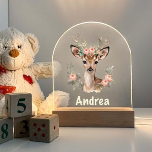 May include: A white acrylic night light with a deer illustration and floral accents. The light has a wooden base and the name "Andrea" is printed below the deer.