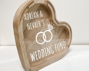 Op de afbeelding: Houten spaarpot in de vorm van een hart met een transparante plastic voorkant. De voorkant heeft een wit ontwerp van twee ringen met een diamant, en de tekst "Adrian & Nevaeh's Wedding Fund".