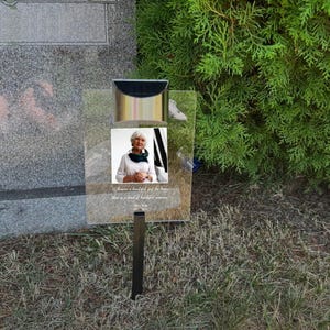 May include: A clear memorial plaque featuring a photograph of a woman, a gold-coloured top, and a black metal stake. The plaque has engraved text and is positioned in front of a headstone and green foliage.