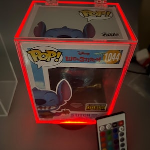 Op de afbeelding: Een rode, verlichte vitrine met een Funko Pop! figuur van Stitch uit de Disney-film Lilo & Stitch. Het figuur is blauw en heeft een glinsterende afwerking. De vitrine heeft een transparante acrylglas voorkant en een zwarte basis. De vitrine is verlicht met rode LED-lampjes. De vitrine heeft ook een afstandsbediening om de kleur van de lampjes te veranderen. De vitrine is gelabeld met "Pop! Disney Lilo & Stitch 1044" en "Exclusive".