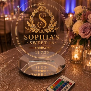 May include: A circular clear acrylic sign featuring gold lettering and a decorative crest with the letter S, the text SOPHIA'S SWEET 16, and the date 11.7.26. The sign is mounted on a round mirrored base and includes a small remote control for lighting.