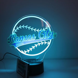 May include: A baseball-themed night light with the words "Kansas City" in a stylized font, and the name "Steve" below. The light is outlined in white and glows with a blue hue. It sits on a black base with a remote control.