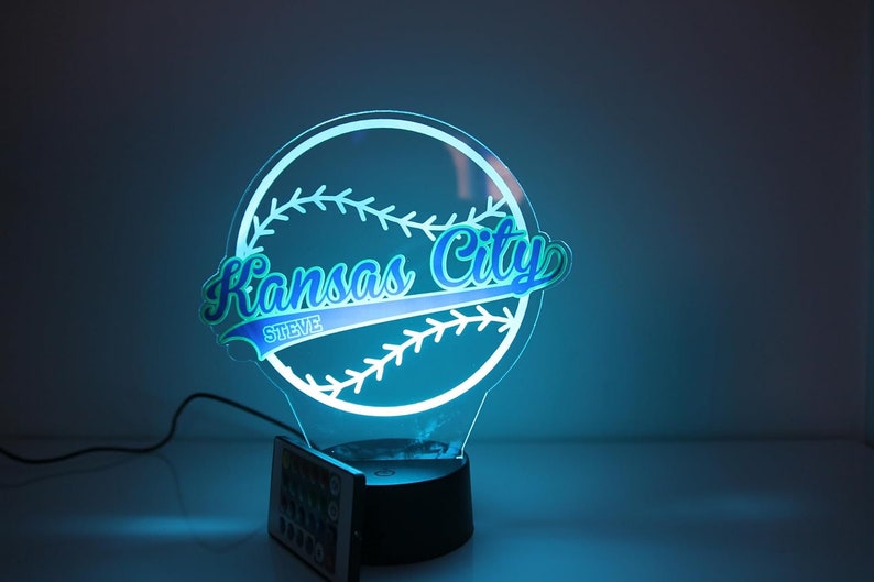 May include: A baseball-themed night light with the words "Kansas City" in a stylized font, and the name "Steve" below. The light is outlined in white and glows with a blue hue. It sits on a black base with a remote control.