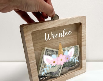 Può includere: Salvadanaio in legno con un fronte trasparente, che presenta un design di unicorno con fiori rosa e blu e un corno dorato. Il nome "Wrenlee" è scritto sul davanti.