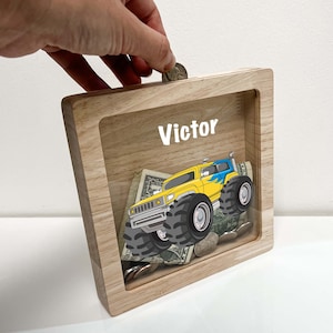 May include: A wooden piggy bank with a clear plastic front. The front features a cartoon monster truck and the name "Victor".