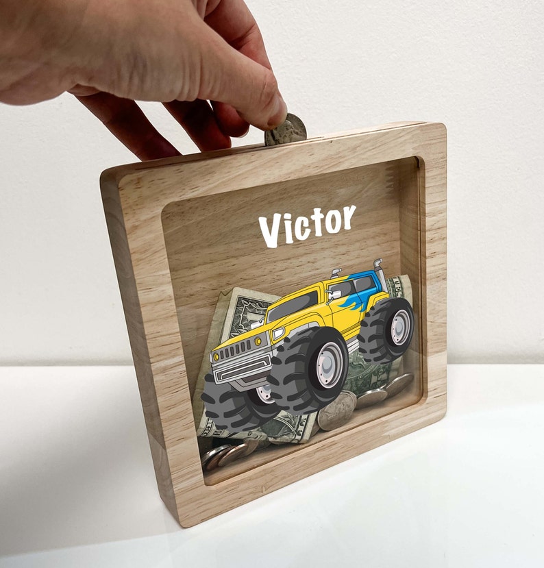 May include: A wooden piggy bank with a clear plastic front. The front features a cartoon monster truck and the name "Victor".