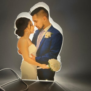 Puede incluir: Una luz nocturna de acrílico personalizada con una pareja en atuendo de boda. La luz se ilumina desde atrás, creando un brillo suave alrededor de la imagen.