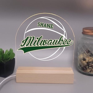 May include: A clear acrylic basketball-themed night light with the name "Shane" and "Milwaukee" in green and white. The light sits on a wooden base and is connected to a white cord. A jar with a copper lid is visible in the background.