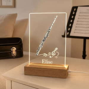 May include: A rectangular acrylic night light with a wooden base, featuring an engraved flute illustration, musical notes, a treble clef, and the name Elise. The light is illuminated with a warm glow and includes a white power cord.