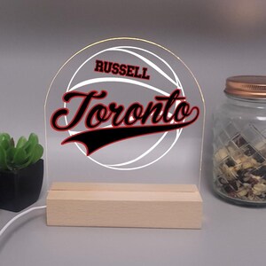 May include: A clear acrylic LED sign with the text "RUSSELL Toronto" in red and white, set against a basketball graphic. The sign is mounted on a wooden base and illuminated with a warm glow. A small plant and a jar are in the background.