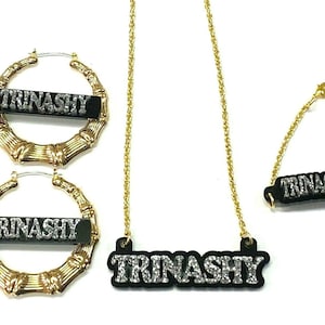 May include: Gold bamboo hoop earrings and a necklace with a black pendant that says "TRINASHY" in silver glitter letters.