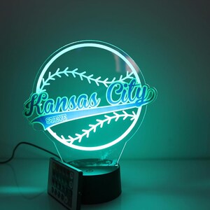 May include: A Kansas City baseball-themed LED lamp. The lamp features a baseball shape with the words "Kansas City" in blue and green, and "STEVE" below. The lamp has a black base and a remote control.