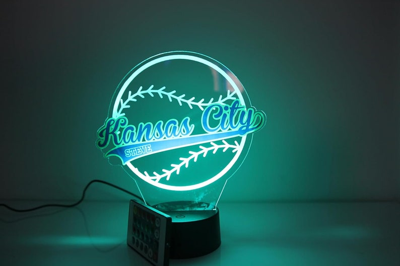 May include: A Kansas City baseball-themed LED lamp. The lamp features a baseball shape with the words "Kansas City" in blue and green, and "STEVE" below. The lamp has a black base and a remote control.