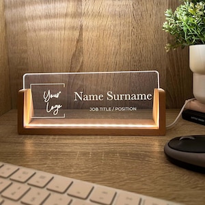 Peut inclure: Plaque de bureau éclairée avec une base en bois. Le panneau en acrylique transparent affiche le texte "Your Logo", "Name Surname" et "JOB TITLE / POSITION". La plaque nominative est éclairée d'une lueur chaude, posée sur un bureau en bois.