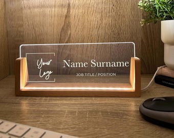 May include: A desk nameplate with a wooden base and illuminated acrylic panel. The text on the panel reads "Your Logo", "Name Surname", and "JOB TITLE / POSITION". The nameplate emits a warm light and is placed on a wooden desk.