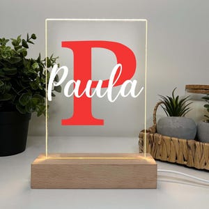 Könnte beinhalten: Personalisiertes LED-Nachtlicht mit Holzsockel. Die transparente Acrylplatte zeigt einen großen roten Buchstaben "P" und den Namen "Paula" in weißer Schrift. Das Licht ist an und beleuchtet den Text.