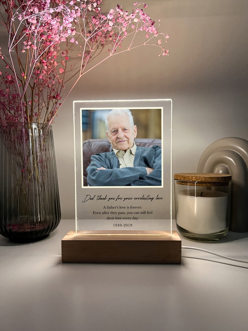 May include: A wooden base with a clear acrylic frame containing a black and white photo of an older man. The frame has a white LED light around the edge. The text "Dad, thank you for your everlasting love. A father's love is forever. Even after they pass, you can still feel their love every day. 1949-2019" is printed on the acrylic frame.