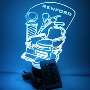 Puede incluir: Lámpara LED azul iluminada con diseño de barbería, que incluye un poste de barbero, tijeras y una silla. El nombre "RENFORD" está en la parte superior. Un mando a distancia y un cable de alimentación son visibles.