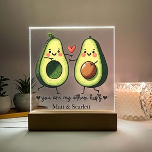 May include: A light-up acrylic sign featuring two cartoon avocados holding hands. The avocados have happy faces and are green with brown pits. The sign reads "You are my other half" with a heart symbol on each side of the text. Below the text is "Matt & Scarlett".