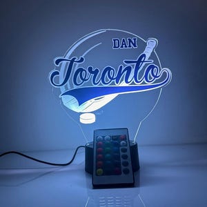 May include: A blue and white illuminated Toronto baseball team logo night light. The light has the name "DAN" above the word "Toronto" in a script font. A remote control is included to change the light's colors.