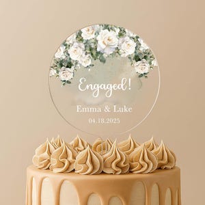 Peut inclure: Un gâteau topper circulaire en acrylique transparent orné d'un arrangement floral aquarelle de roses blanches et de feuilles vertes sur le dessus. Le texte indique Engaged! suivi des noms Emma & Luke et de la date 04.18.2025 en blanc.