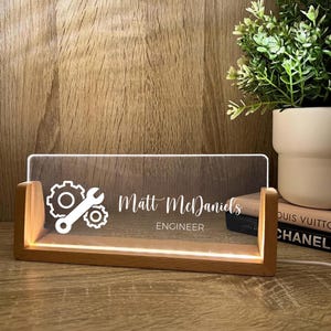 Puede incluir: Placa de escritorio personalizada para un ingeniero. La placa de acrílico transparente está iluminada por una luz cálida y presenta el nombre "Matt McDaniels" y la palabra "ENGINEER" en letras blancas, junto con gráficos de engranajes y llaves.