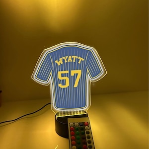 May include: A blue and yellow illuminated night light shaped like a baseball jersey. The jersey has the name "WYATT" and the number "57" on the back. A remote control is in the foreground.